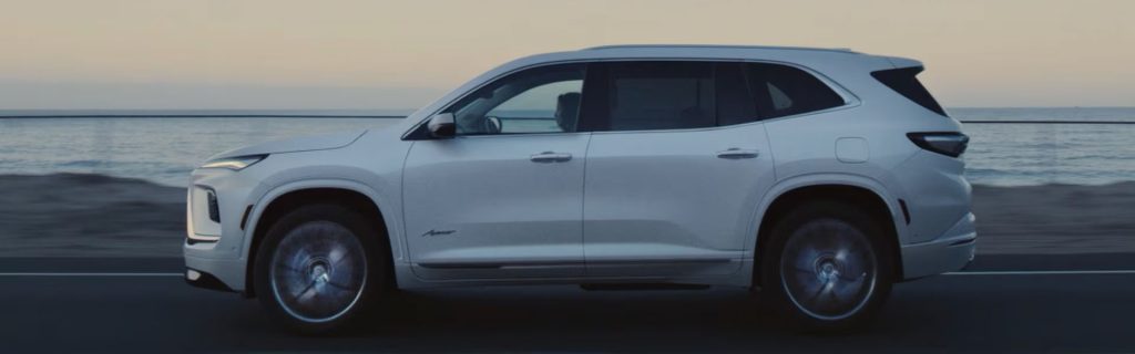 A white Buick Enclave driving along a coastal road at sunset