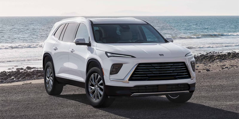 A white Buick Enclave parked on a paved area along a sandy beach with the ocean waves in the background.