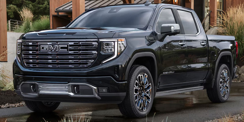 Front-three-quarter view of a black GMC Sierra Denali Ultimate parked on a wet pavement in front of a modern wood-accented building.