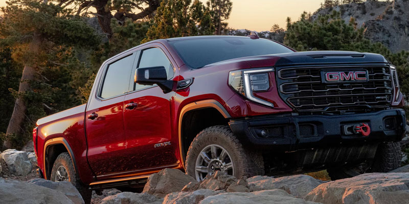 Front-three-quarter view of a dark red GMC Sierra truck navigating a rocky off-road terrain at sunset.
