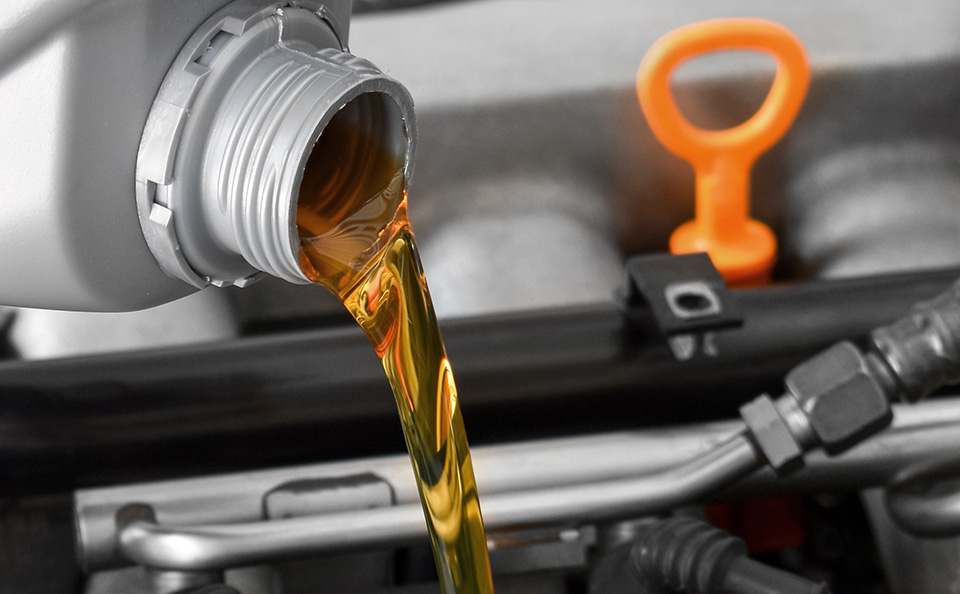 Close-up of amber-colored fresh engine oil being poured into a car engine with an orange dipstick handle visible in the background.