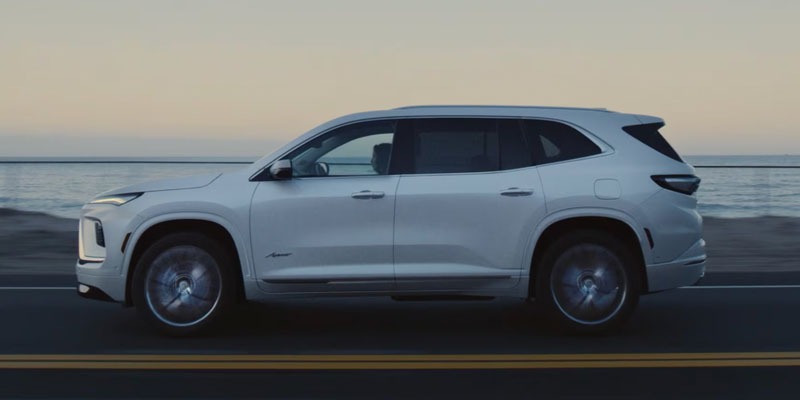 Side view of a white Buick Enclave driving along a coastal highway with the ocean in the background during twilight.