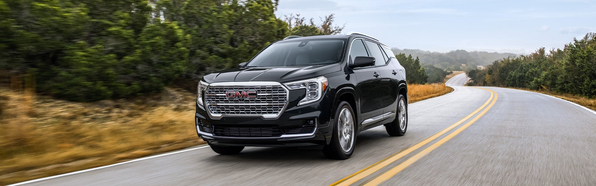 A pre-owned black GMC Terrain compact SUV driving along a winding paved road next to lush green trees.
