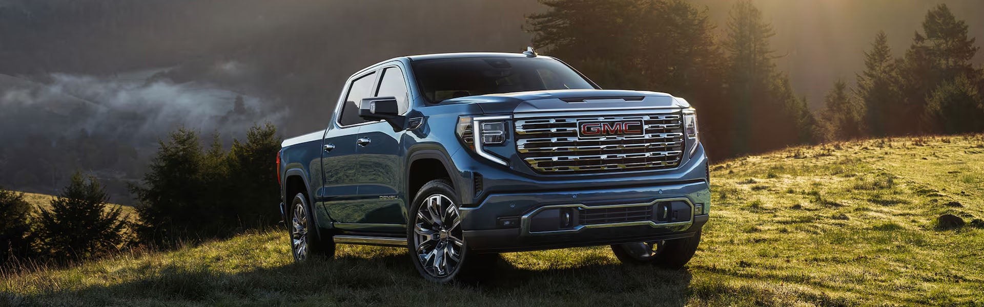A dark blue GMC Sierra 1500 pickup truck parked on a grassy hill surrounded by evergreen trees at sunrise.