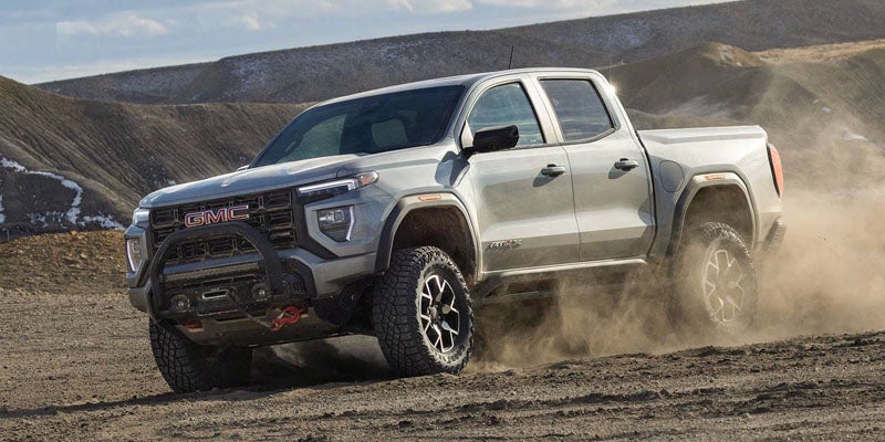 A silver GMC Canyon AT4X pickup truck driving off-road through dirt and kick-up dust.