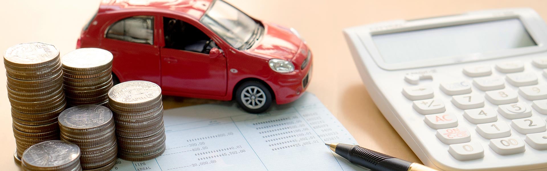 A red toy car sitting on a bank passbook next to stacks of coins, a pen, and a white calculator on a desk, representing auto financing or budgeting.