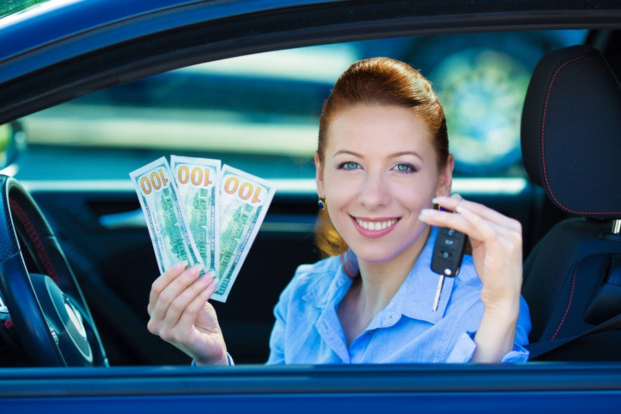 A smiling woman sitting inside a car holding a stack of cash bills and a car key fob.